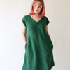 Rae is wearing a green v-neck dress with a curved hem. She has her hands in her pockets and wears glasses. She looks off to the side. 