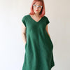 Rae is wearing a green v-neck dress with a curved hem. She has her hands in her pockets and wears glasses. She looks off to the side.