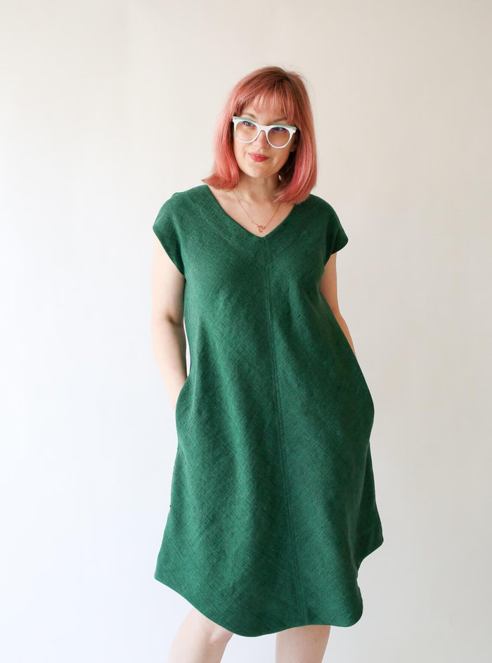 Rae is wearing a green v-neck dress with a curved hem. She has her hands in her pockets and wears glasses. She looks off to the side.