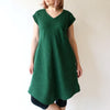 Rae is wearing a green v-neck dress with a curved hem. She has her hands at her side. She looks off to the side.